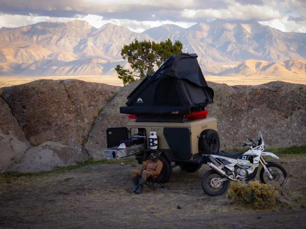 Bean Pod at camp with a rooftop tent and kayaks, a dirt bike parked beside it