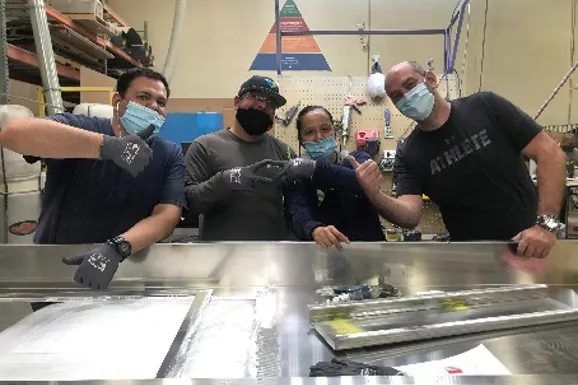 Four Bean team members in masks giving a thumbs up around a steel work table in the plant