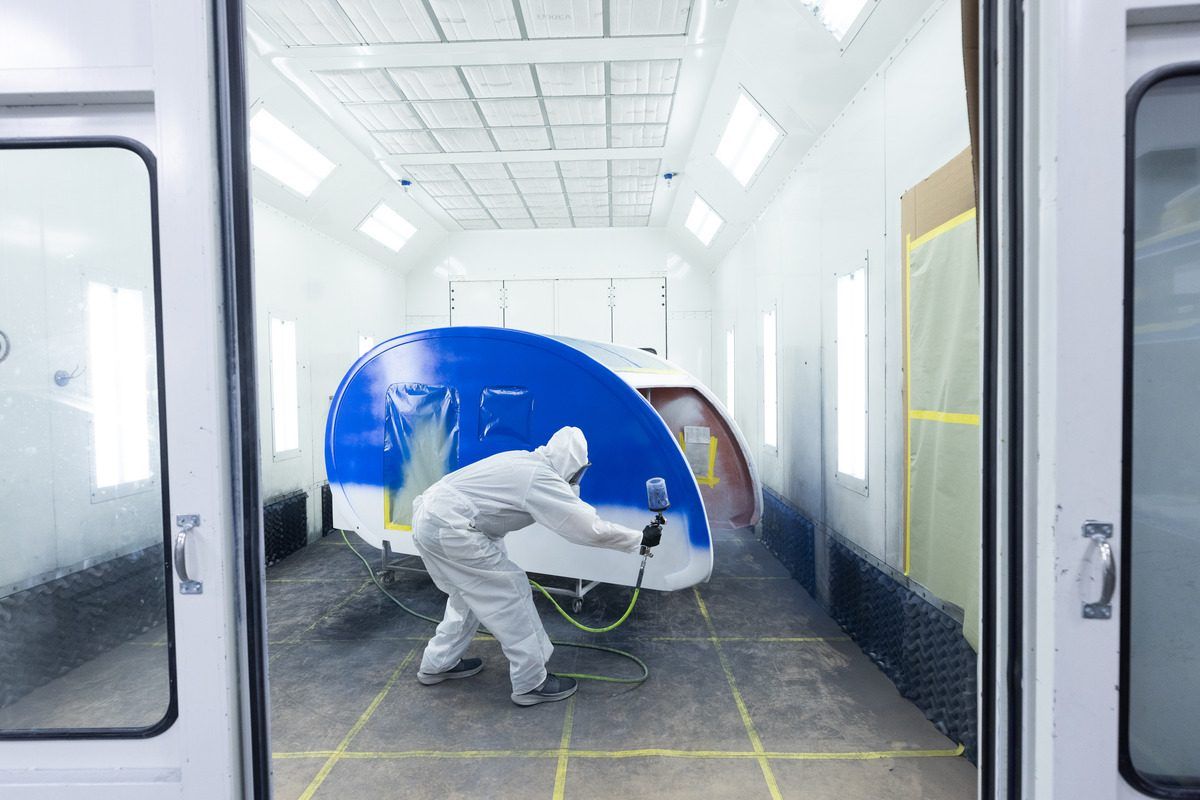 A technician in white coveralls painting a blue fiberglass shell in the paint booth
