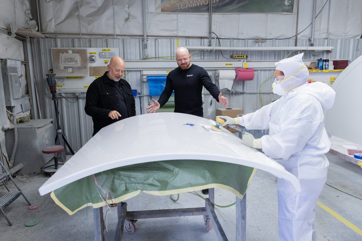 Mark and Brian beside a technician sanding a white fiberglass shell in the plant