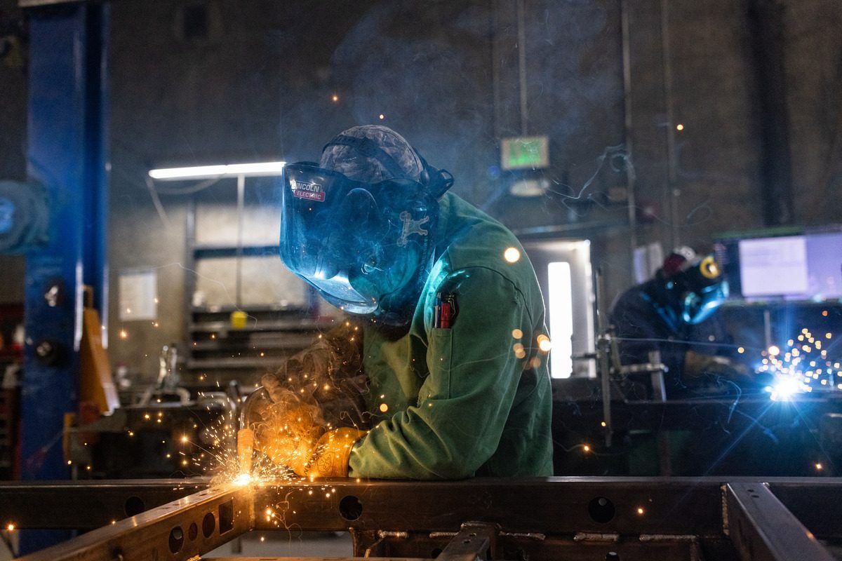 A welder in a green jacket and helmet welding a steel frame, sparks flying